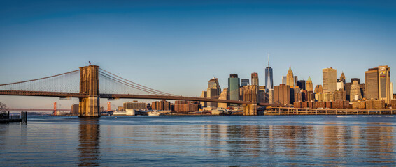 Fototapeta premium Iconic Brooklyn Bridge and Lower Manhattan Skyline at Golden Hour, New York City. Panoramic view of historic architecture, skyscrapers, and East River reflections under a clear blue sky.
