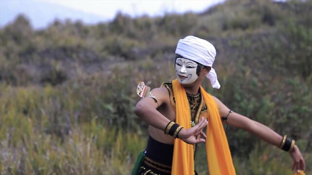 Traditional Indonesian dancer in a white mask and yellow sash performing on a grassy hill, 4K video.