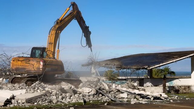 Demolition excavator and hydraulic breaker breaking a concrete structure on the beach. Wide view, sunny day, blue sky and sea on background