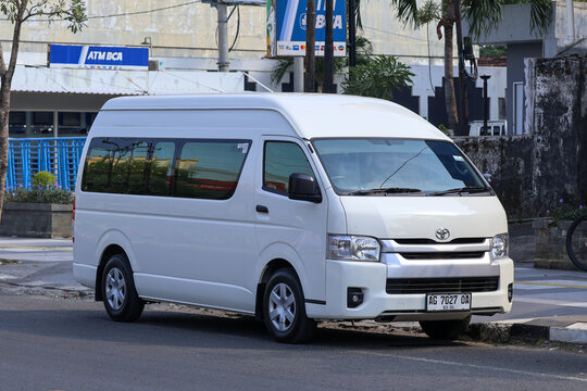 A white Toyota HiAce commercial commuter van parked on the side of a street with shop signs and buildings in the background.