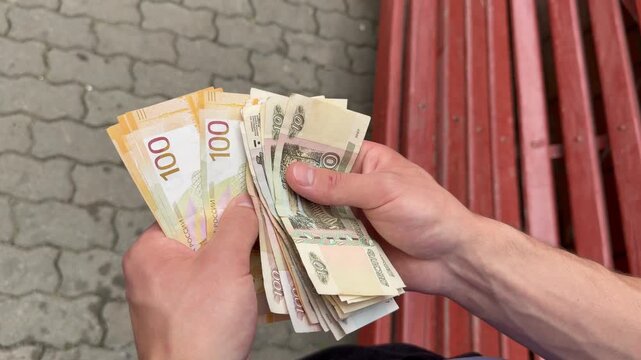 Close up view of a man counting Russian ruble banknotes outdoors sitting on a wooden bench, including new 100 ruble bills. A man counts Russian money on the blurred background. 
