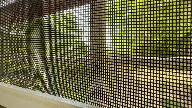 A dark mesh screen covers the window. It filters the bright view of a green summer garden outside. The peaceful scene brings a feeling of calm and cozy longing.