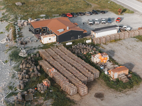 Aerial view of a commercial fishing facility with rows of stacked crab pots, a building with a brown roof, and parked pickup trucks in a gravel lot in Unalaska, Alaska, United States.