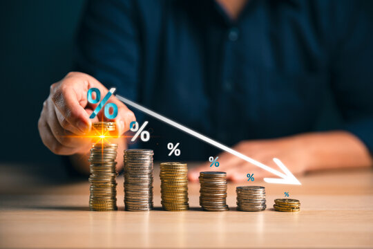 Hand placing coins on stacked money with percent symbols and a downward arrow shows falling interest rates, financial decline, market downturn, savings risk, loan costs and economic crisis concept.