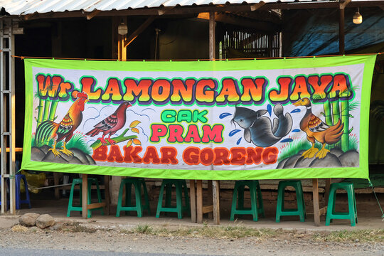 A traditional hand-painted fabric banner of a local street food stall known as "Warung Tenda Pecel Lele Lamongan Jaya Cak Pram", displaying colorful illustrations of a rooster, bird, catfish, and duck