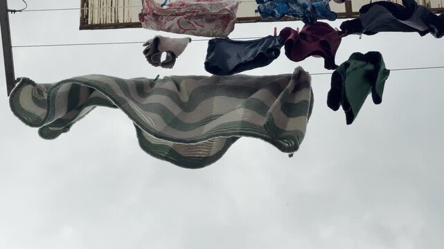Multiple garments and a striped blanket hang to dry on outdoor clotheslines. The items sway gently under a dull, overcast sky. The scene feels quiet, domestic, and simple.