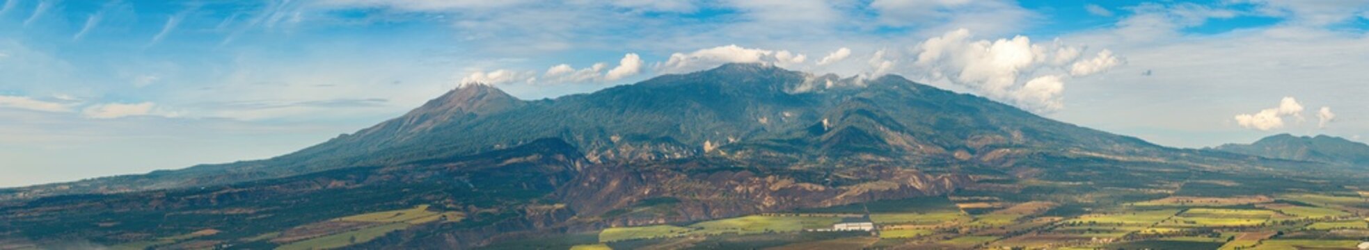 Aerial view of Volcan de Colima rising above lush green slopes and agricultural fields under a bright blue sky with scattered clouds in Jalisco, Mexico.