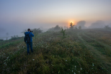 A photographer with a backpack stands in a grassy field, capturing a misty sunrise that casts a soft, golden glow © Александр Арендарь