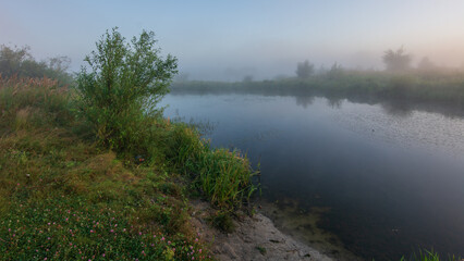 Obraz premium A serene, misty morning landscape features a calm river reflecting the soft sky, surrounded by lush green banks and hazy trees