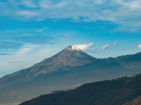 Aerial view of Volcan de Colima with snow-capped peak and surrounding forested slopes under a blue sky in Jalisco, Mexico.