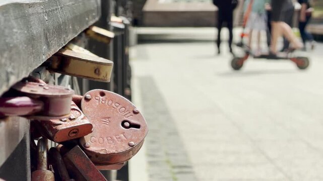 A rusty heart-shaped wedding lock clings to a metal railing. Blurry people walk along the sunny path in the background. This sweet tradition evokes a deep feeling of romantic nostalgia.