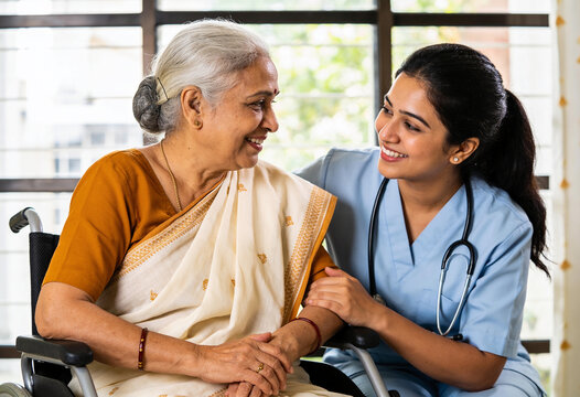 Smiling Indian female nurse assisting elderly senior woman in wheelchair at home bright natural light care