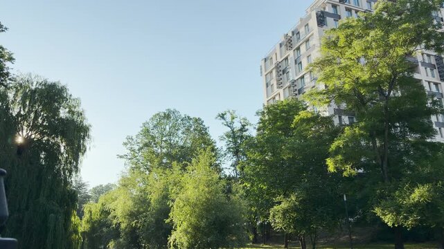 A modern apartment building rises behind a lush green park in Simferopol, Crimea. Bright morning sunlight creates a fresh, peaceful, and cheerful atmosphere under the clear blue sky.