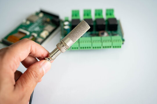 Close-up of an industrial temperature and humidity sensor held by hand with electronic controller board in the background, representing IoT, environmental monitoring, smart automation systems.