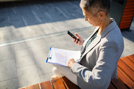 Senior businesswoman using smartphone and reviewing documents on outdoor bench