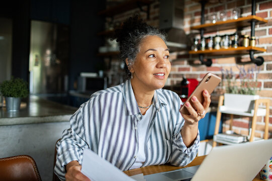 Mature woman using smartphone voice assistant while working in home kitchen