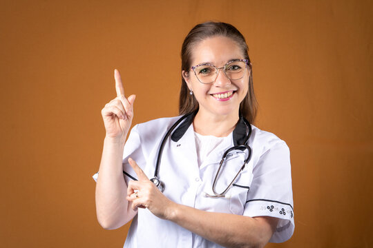 Mujer doctora con lentes, ambo, y estetoscopio se&ntilde;alando hacia arriba con expresi&oacute;n de confianza y seguridad..