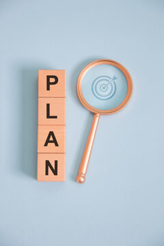 Business planning concept with wooden letter blocks spelling PLAN and magnifying glass highlighting target strategy on blue background, symbolizing goal setting, project management, vision, success.