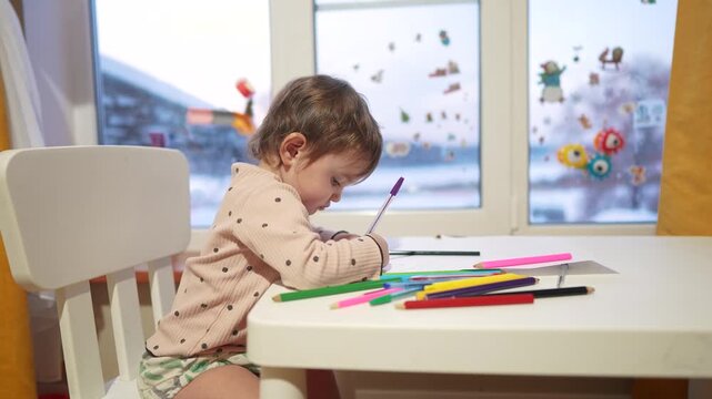 baby girl draws while sitting at table by the window at home. happy family kid dream concept. baby daughter learns to draw with pencils on a sheet of paper indoors. development of fine motor skills