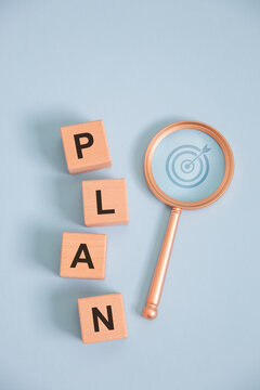 Business planning concept with wooden letter blocks spelling PLAN and magnifying glass highlighting target strategy on blue background, symbolizing goal setting, project management, vision, success.