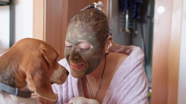 mature woman wearing a cosmetic clay mask and a bathrobe spending quality time with her beagle dog at home, showing affection and enjoying a relaxing beauty treatment routine in the bathroom