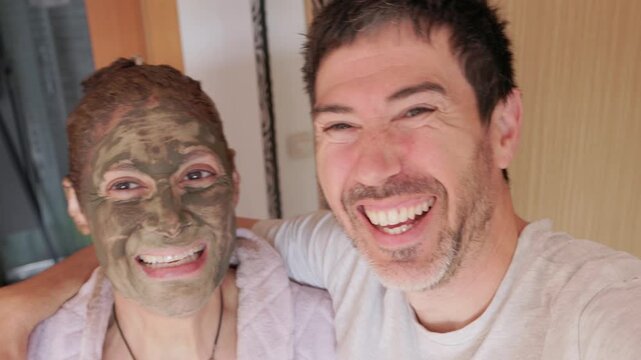 Funny couple taking a selfie while the woman wears a green clay mask for skincare treatment at home. The man and woman are laughing together, enjoying a moment of complicity and fun. ugc content.