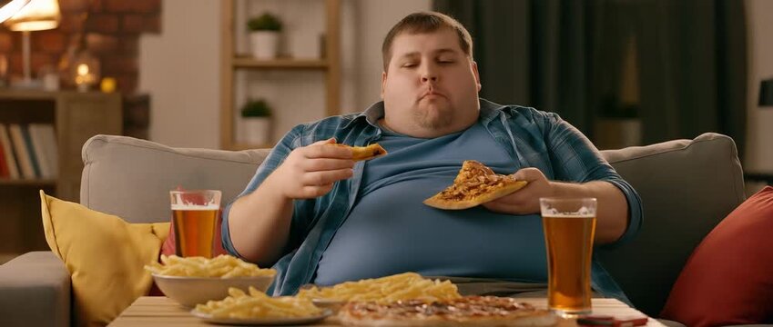 Overweight Man Eating Fast Food on Sofa, represents unhealthy eating habits
