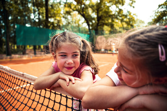 Two young girls leaning on tennis net at outdoor clay court