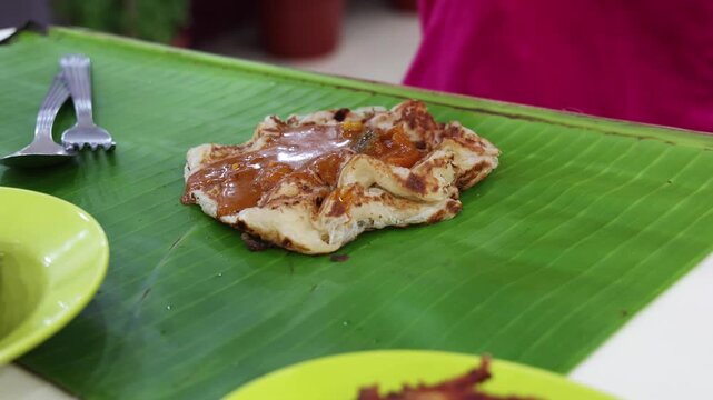Hand with ladle pouring curry onto roti canai served on banana leaf in Indian restaurant in Malaysia. Roti canai is popular breakfast among local Malaysians.
