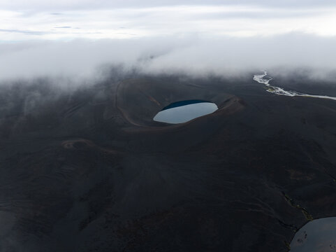 Aerial view of a volcanic crater lake surrounded by dark volcanic soil and low-hanging clouds in Iceland.