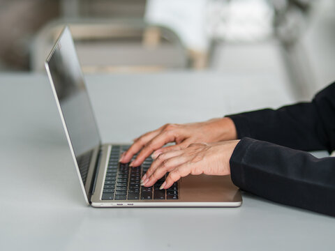 Side view close-up of a businesswoman in a formal black suit typing on a laptop computer keyboard on a white table, focusing on corporate project management.