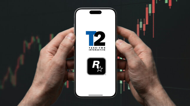 Take-Two Interactive T2 and Rockstar Games Logos Displayed on Smartphone Screen Held with Both Hands Against Stock Market Candlestick Chart Background
