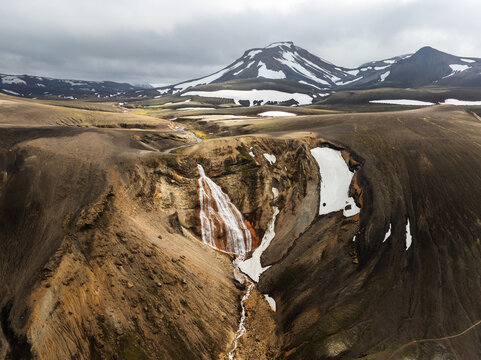 Aerial view of a waterfall cascading down a rugged, volcanic landscape with patches of snow and dark, textured mountains in the Highlands of Iceland.