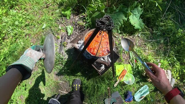 POV of man opening lid and stirring hot goulash soup in cauldron over campfire