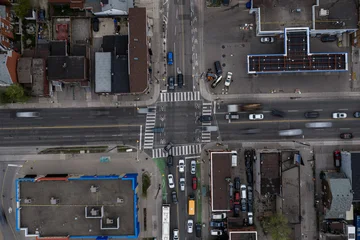 Aerial top down view of busy Toronto city intersection with moving cars, marked crosswalks, bike lanes, rooftops, parking areas, and dense urban blocks under overcast light © 21AERIALS