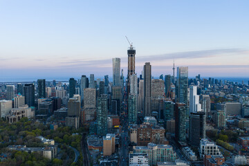 Obraz premium Toronto skyline at dusk with high rise towers, construction crane, distant lake horizon, layered downtown buildings, soft evening light, clear elevated city view over urban core