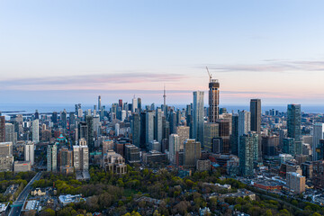 Obraz premium Toronto skyline at dusk with dense downtown towers, CN Tower, rising high-rises, construction crane, lake horizon, pastel evening sky, urban neighborhoods, and tree-lined foreground