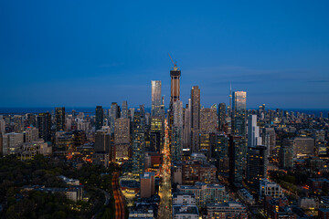 Obraz premium Dusk aerial view over Toronto skyline with illuminated high-rise towers, dense downtown streets, and bright blue evening sky stretching above Lake Ontario