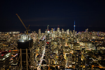 Obraz premium Night aerial view over downtown Toronto with illuminated high rise towers, construction crane on high rise building foreground, dense street grid, and tall landmark tower glowing against dark sky
