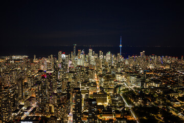 Obraz premium Night aerial view of Toronto skyline with illuminated towers, CN Tower rising above dense downtown buildings, dark lake horizon, glowing city grid and urban lights