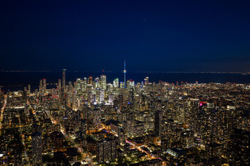 Obraz premium Toronto night skyline glowing above dense downtown towers with city lights, illuminated streets, distant lake horizon, and iconic central tower seen from high aerial viewpoint