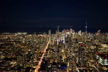 Obraz premium Toronto skyline glowing at night with dense downtown towers, illuminated streets, dark lake horizon, and iconic city lights viewed from high aerial perspective over Canada