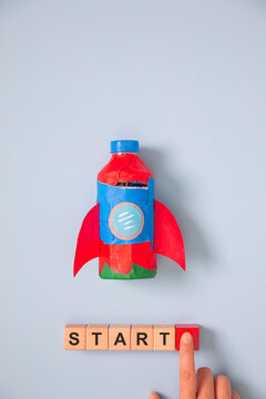 Creative startup concept with handmade rocket, wooden START blocks and finger pressing launch button on blue background, symbolizing business growth, innovation, entrepreneurship, motivation, success.