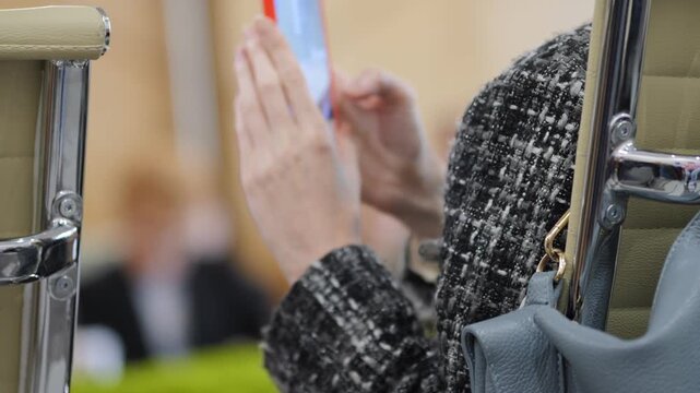 Person in a tweed jacket uses a smartphone while seated. They hold the device with both hands, focused on the screen. The background shows blurred meeting attendees. This shot works well for business