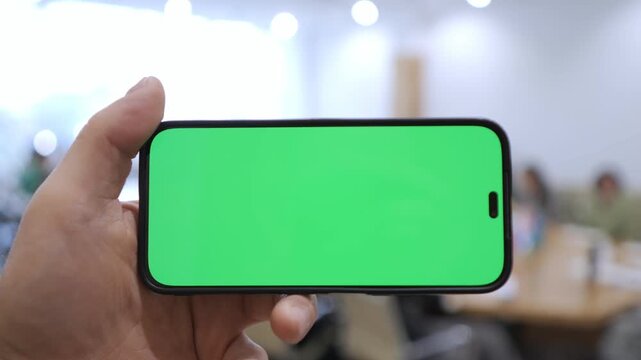 Hand holds phone with green screen. Background shows office space. Phone is held steady for overlays. Perfect for app or website mockups. Green screen allows easy editing