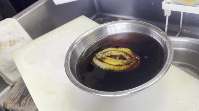 A chef pours soy sauce into a bowl with a cleaned abalone and kombu kelp, preparing steamed abalone in a Japanese kitchen.