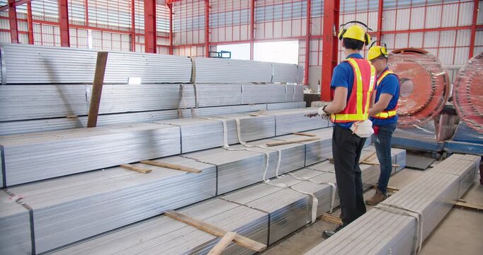 Two factory workers inspect metal construction materials in a warehouse, performing quality control and managing the industrial supply chain and inventory.