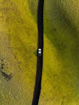 Aerial view of a white car driving on a dark gravel road through vibrant green mossy volcanic fields near Laki in Skaftarhreppur, Iceland.