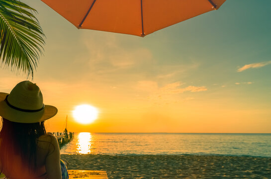 Back view of a woman in a bikini relaxing beneath a beach umbrella while watching a glowing tropical sunset over calm ocean. Luxury summer vacation, seaside leisure, wellness, and peaceful coastal.