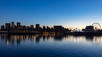 Fototapeta premium Montreal city skyline at dusk seen from the old port waterfront with a ferris wheel, festival tent and calm river reflections, Quebec, Canada